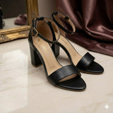 Black high-heeled sandals on a light surface with a mirror and fabric in the background